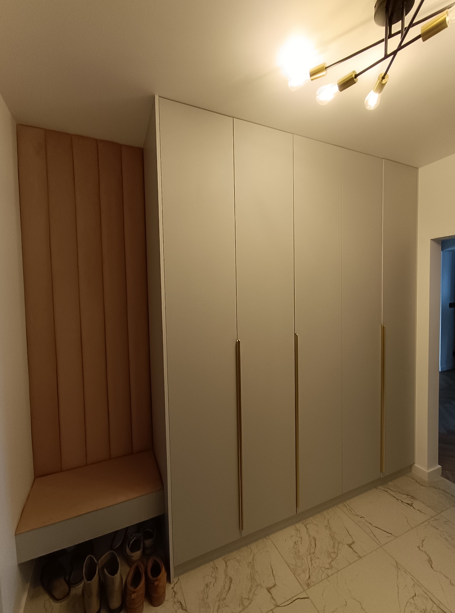 Szafa wnękowa z siedziskiem i panelem tapicerowanym w przedpokoju. Jasne fronty, złote uchwyty, marmurowa podłoga. Pod siedziskiem widoczne buty.