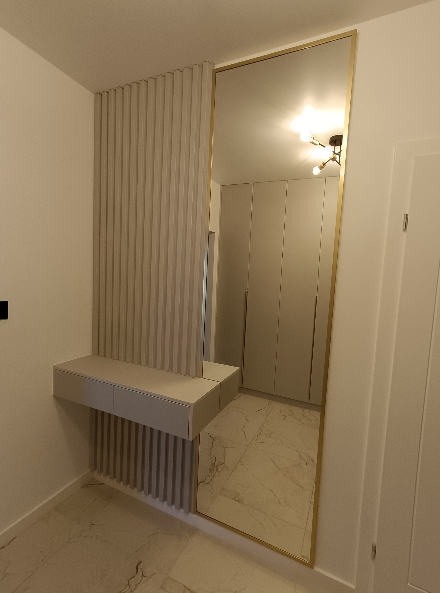 Nowoczesna zabudowa przedpokoju z lustrem w złotej ramie i konsolą z lamelami w odcieniach beżu. Widoczne szafy w tle i marmurowa posadzka.