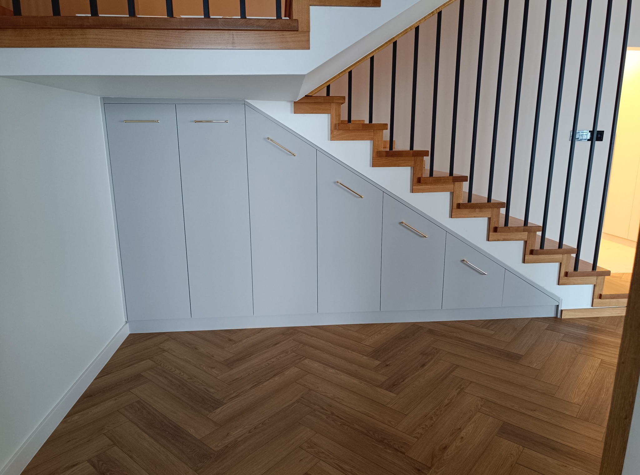 Szafa wnękowa pod schodami z szarymi frontami i złotymi uchwytami, parkiet w jodełkę. Zabudowa wnęki pod schodami.