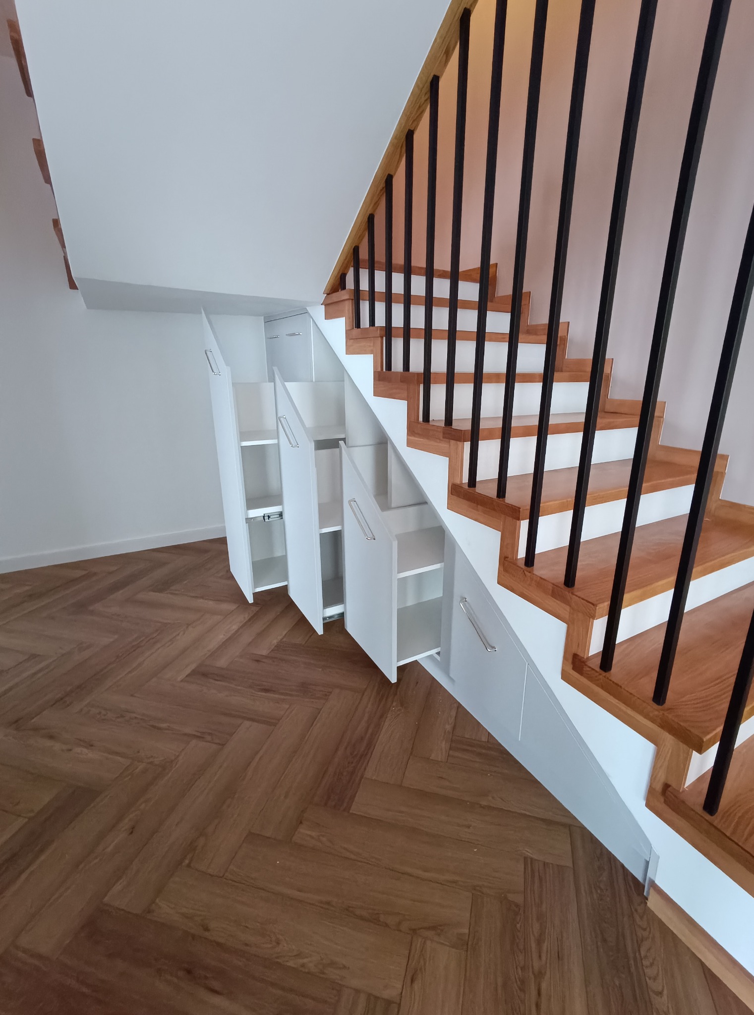Schody z drewnianymi stopniami i czarną balustradą, zabudowane białymi szafkami z uchwytami, otwartymi w połowie, na podłodze parkiet w jodełkę.