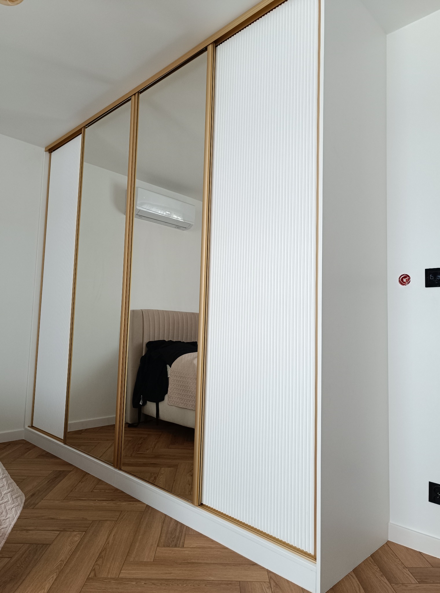 Nowoczesna szafa wnękowa z lustrzanymi drzwiami przesuwnymi i białymi panelami dekoracyjnymi, odbijająca fragment sypialni z łóżkiem i klimatyzacją.