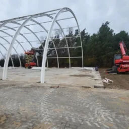 Konstrukcja stalowa hali w trakcie montażu. Widoczny dźwig teleskopowy Manitou i podnośnik koszowy. Na pierwszym planie ułożona kostka brukowa.