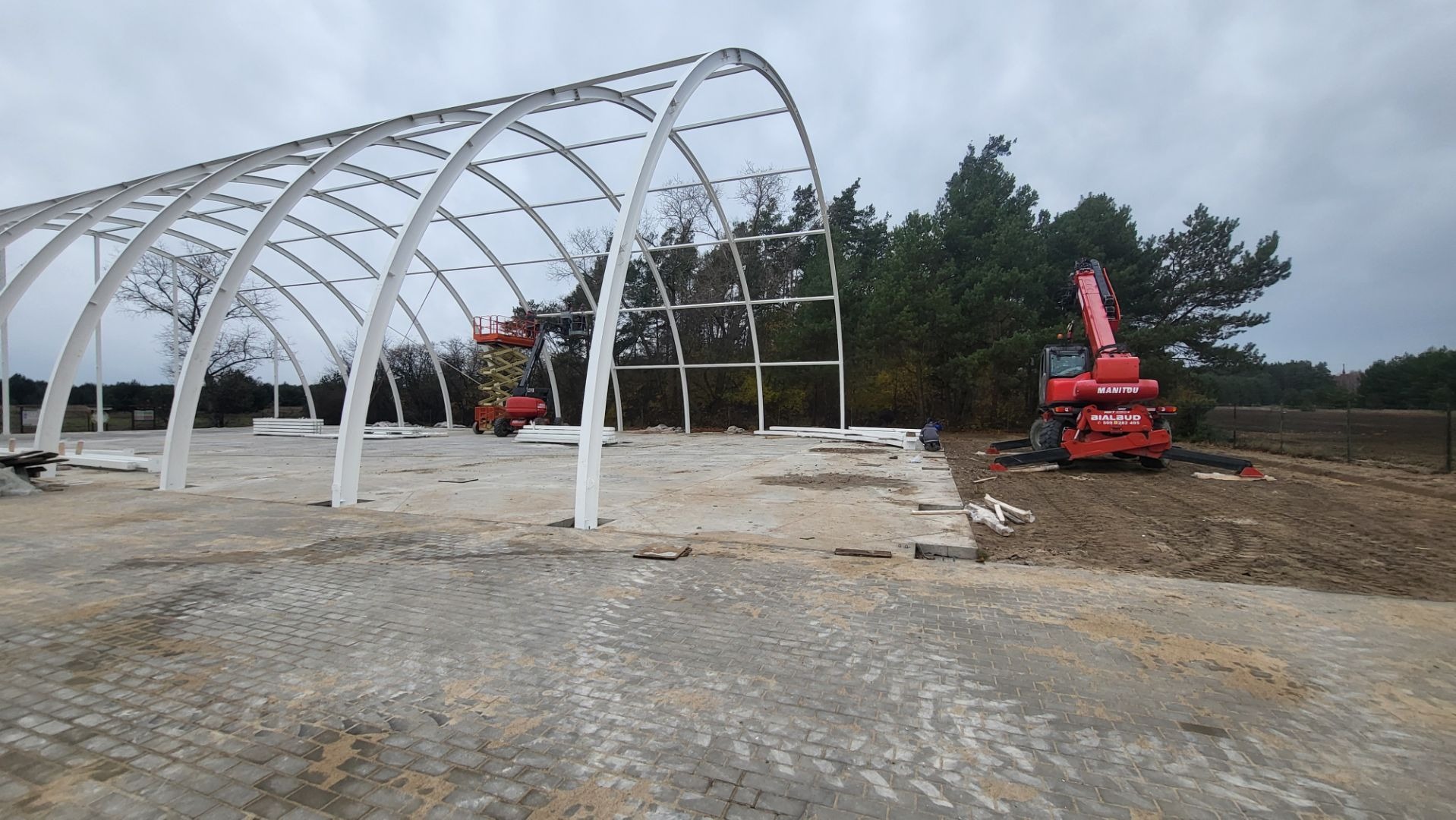 Konstrukcja stalowa hali w trakcie montażu. Widoczny dźwig teleskopowy Manitou i podnośnik koszowy. Na pierwszym planie ułożona kostka brukowa.