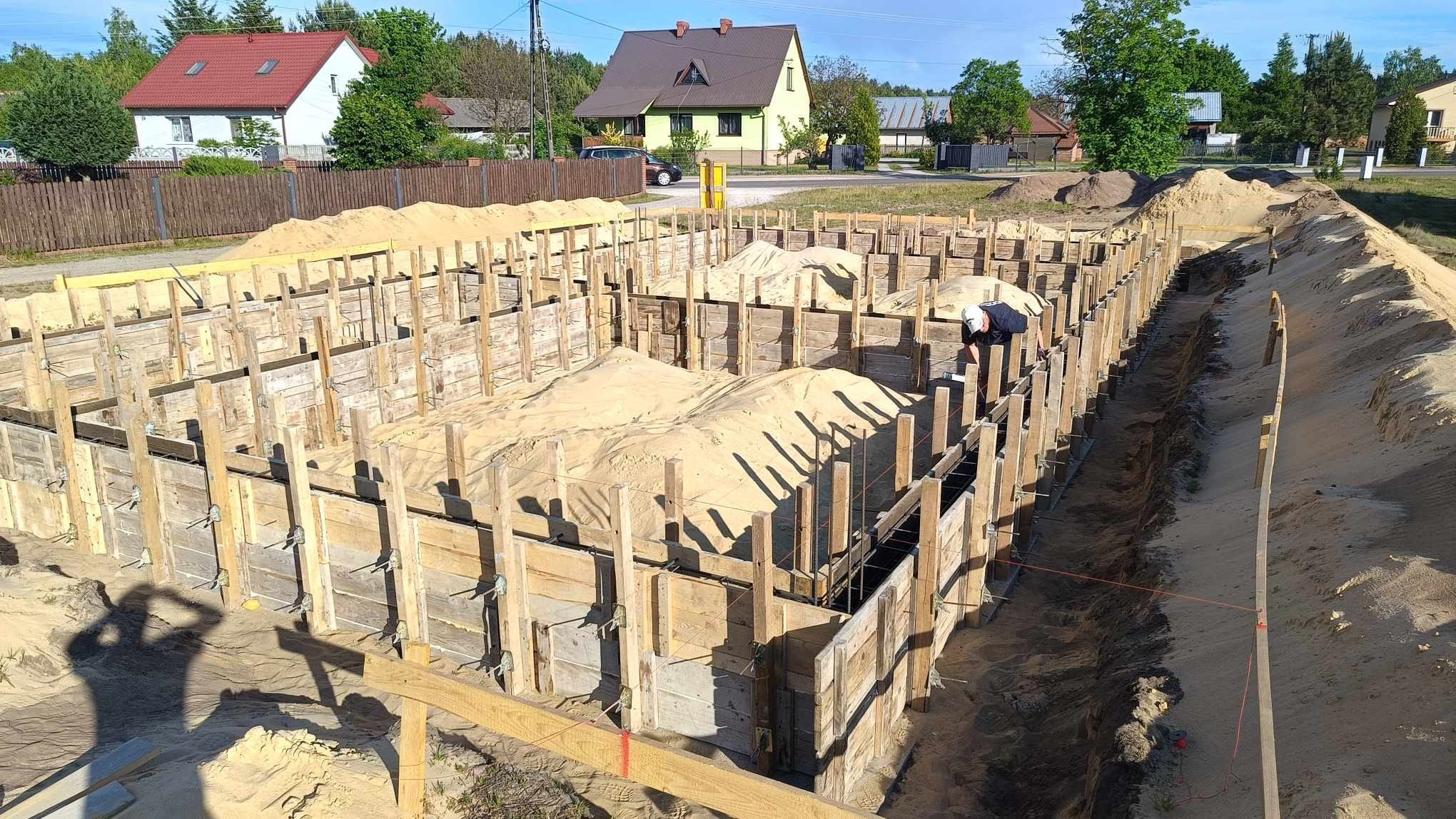Fundamenty domu jednorodzinnego w trakcie budowy: drewniane szalunki, zbrojenie i nasypany piasek. W tle zabudowa mieszkalna i zieleń.