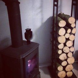 Adrian Pietraniec - Żeliwny piec z płomieniem i stojak z drewnem opałowym w pomieszczeniu.  Dekoracyjny lampion na piecu. Oświetlenie boczne tworzy cień na ścianie.