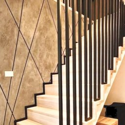 Adrian Pietraniec - Nowoczesne schody z drewnianymi stopniami i czarną, metalową balustradą o geometrycznym wzorze, na tle ściany z dekoracyjnym tynkiem w odcieniach szarości.