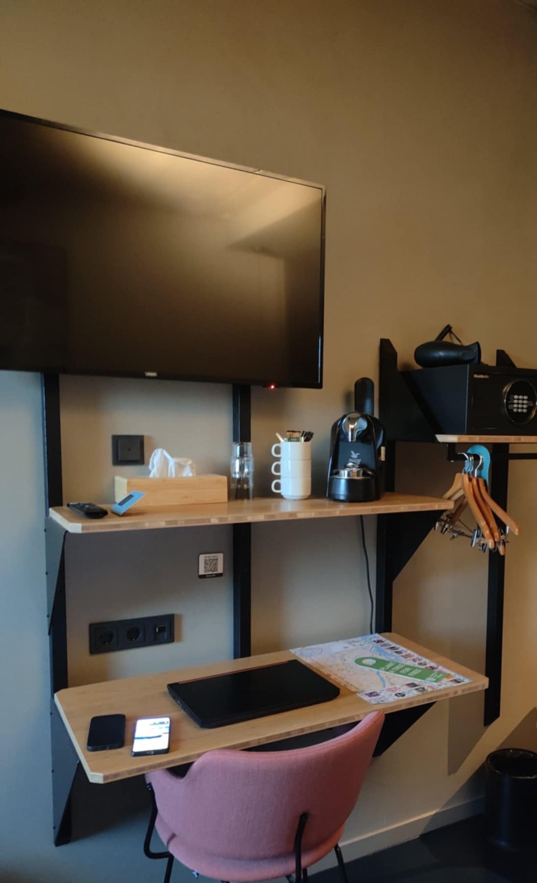 Nowoczesny kącik hotelowy: telewizor, biurko z laptopem, telefonami, ekspres do kawy i sejf, tworzący funkcjonalną przestrzeń w stonowanej kolorystyce.