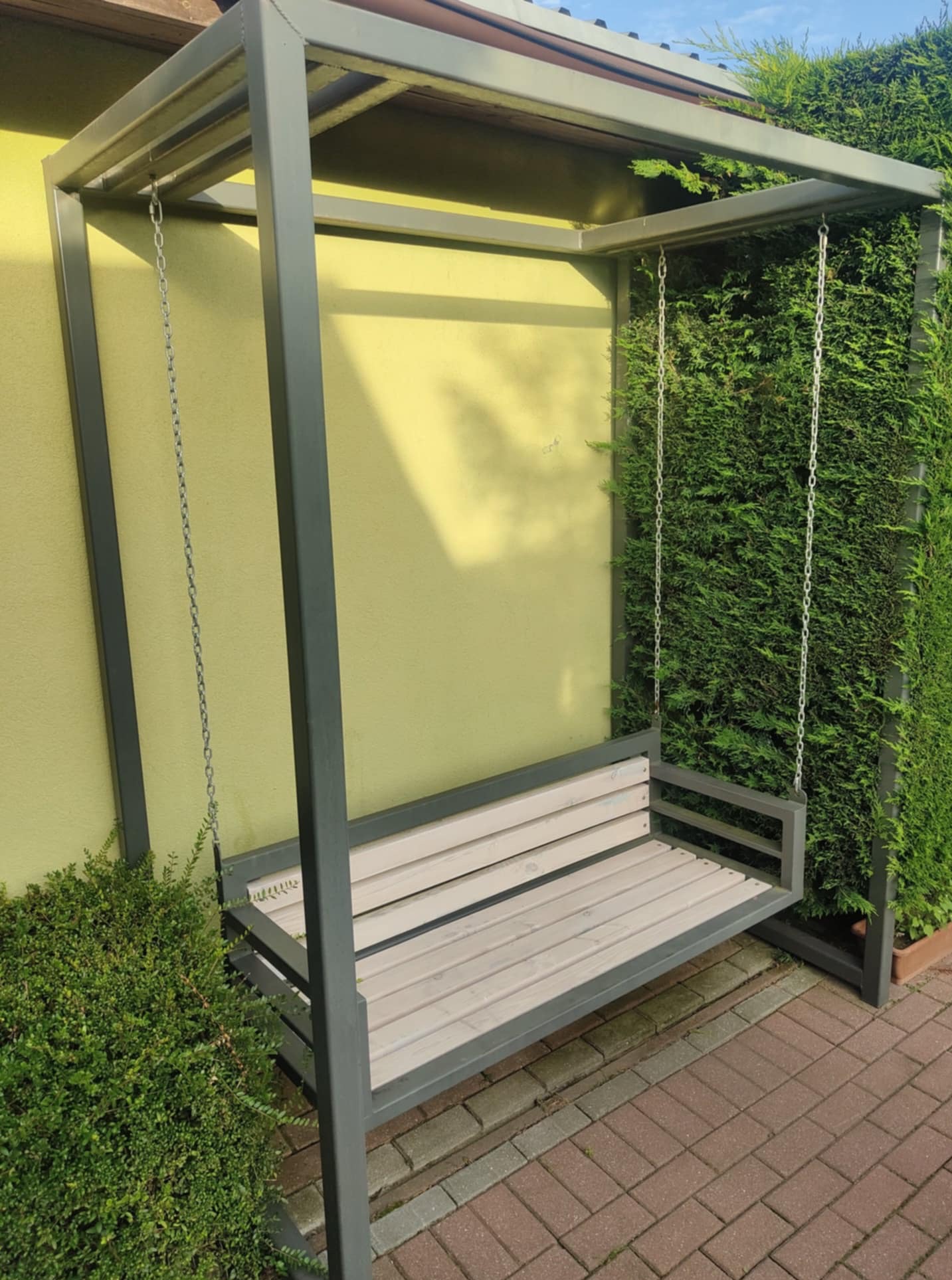 Metalowa huśtawka ogrodowa z drewnianym siedziskiem, zawieszona na łańcuchach, na tle żółtej ściany i zielonego żywopłotu. Widok z boku, kostka brukowa.