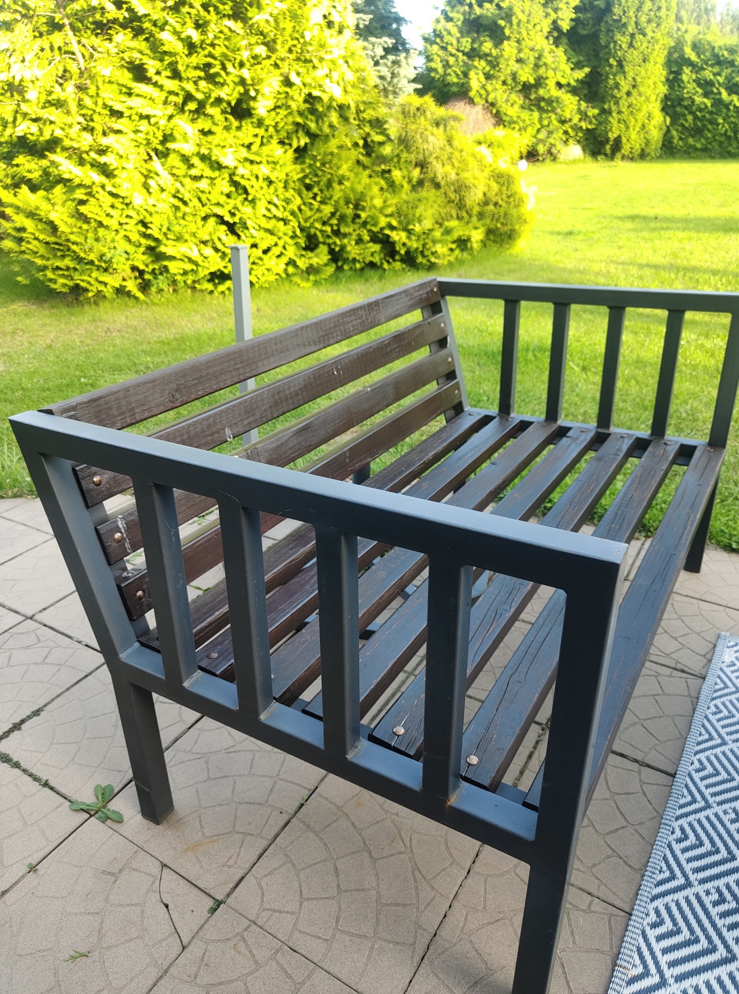 Nowoczesna ławka ogrodowa z ciemnego drewna i metalu na tarasie z kostki, otoczona zielenią. Widok z góry, dywanik z geometrycznym wzorem.