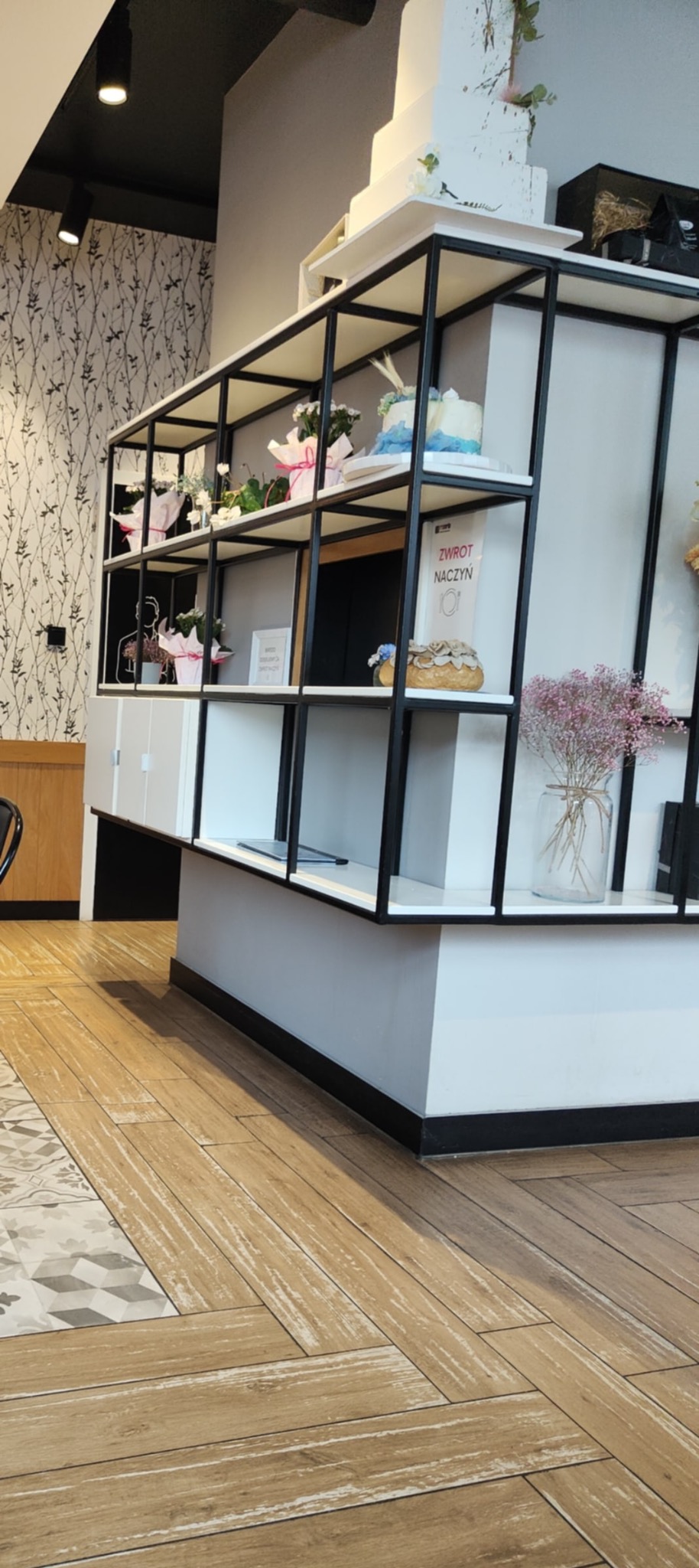 Kawiarnia w Mrzezinie. Ekspozycja tortów i dekoracji kwiatowych na czarno-białym regale, widok z perspektywy podłogi z drewnianych paneli.