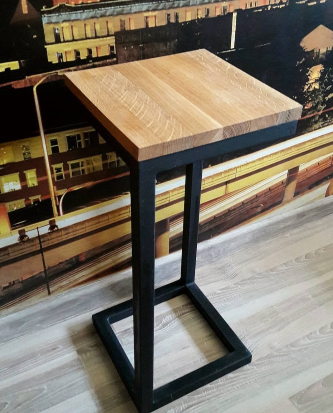 Stolik kawowy z blatem dębowym i czarną, metalową podstawą w kształcie litery C, na tle fototapety z motywem miejskim.