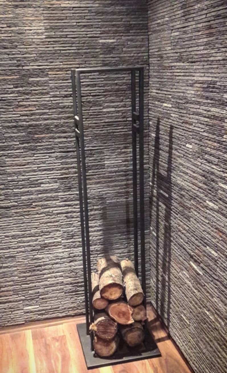 Metalowy stojak na drewno kominkowe w rogu pomieszczenia z kamienną okładziną ściany, drewniana podłoga. Minimalistyczny design.