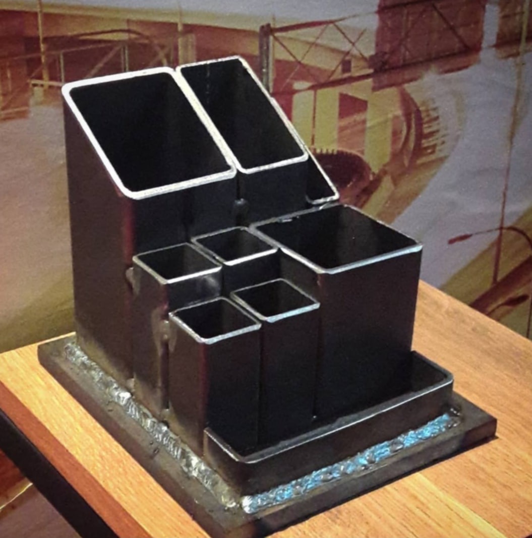 Metalowy organizer biurkowy spawany, z kwadratowych profili, na drewnianym blacie. Widoczne spawy. W tle grafika.