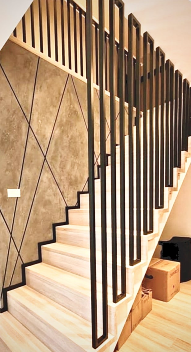 Nowoczesne schody z drewnianymi stopniami i czarną, metalową balustradą o geometrycznym wzorze, na tle ściany z dekoracyjnym tynkiem w odcieniach szarości.