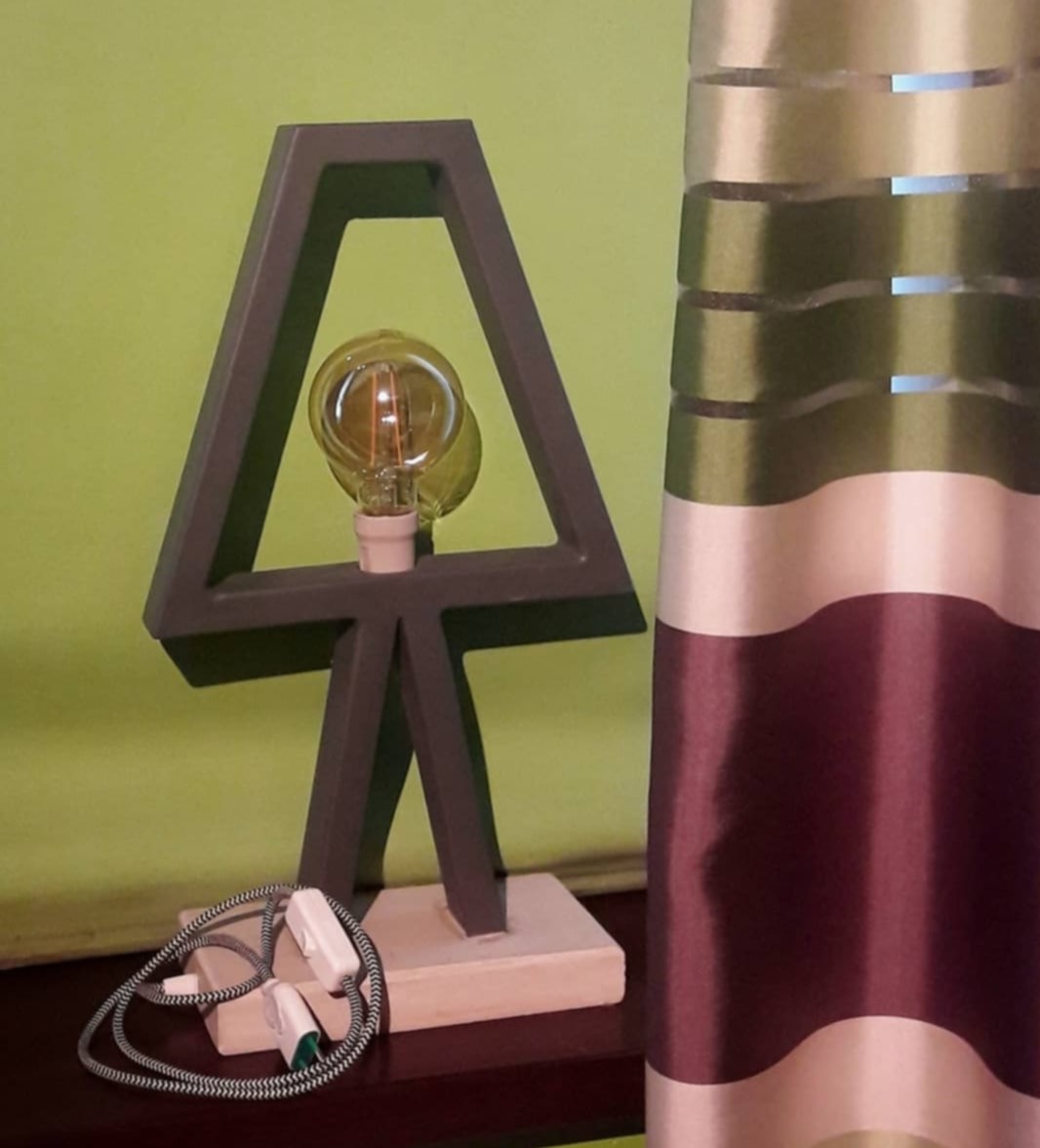 Geometryczna lampka stołowa z żarówką Edisona na tle zielonej ściany i zasłony w pasy. Kabel z włącznikiem leży obok podstawy.