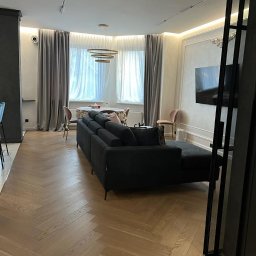 Ruzop - Elegancki salon z parkietem w jodełkę, ciemną sofą i dekoracyjnymi zasłonami. Nowoczesne oświetlenie i telewizor na ścianie. Stonowana kolorystyka, jasne ściany.