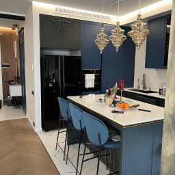 Ruzop - Elegancka kuchnia z wyspą, marmurowym blatem i niebieskimi szafkami. Nad wyspą wiszą trzy ozdobne, szklane lampy. Podłoga wyłożona parkietem w jodełkę.
