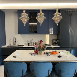 Ruzop - Nowoczesna kuchnia w odcieniach granatu i bieli z marmurowym blatem wyspowym, oświetlona trzema designerskimi lampami wiszącymi. Widoczne detale i dodatki.