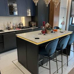 Ruzop - Niebieska kuchnia z wyspą oświetloną listwą LED i marmurowym blatem. Nad wyspą wiszą trzy szklane lampy. Marmurowe płytki na podłodze z akcentami w kolorze złotym.