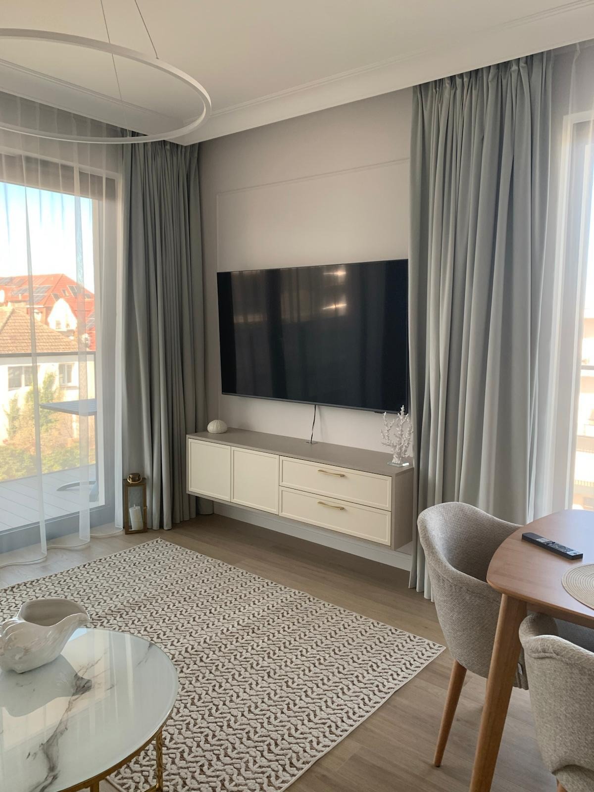 Nowoczesny salon z telewizorem na ścianie, beżową szafką RTV, wzorzystym dywanem i szarymi zasłonami. Widok na balkon przez okno z delikatną firanką.