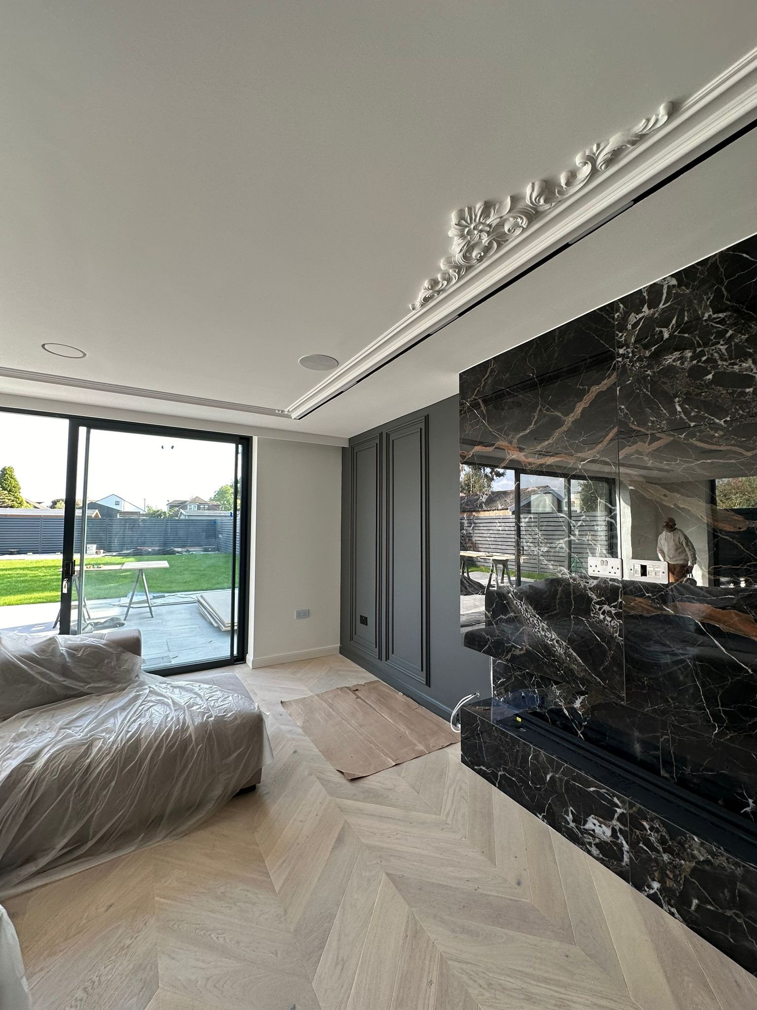 Salon w trakcie wykańczania: parkiet w jodełkę, marmurowy kominek, sofa zabezpieczona folią, duże okno z widokiem na ogród. Elementy dekoracyjne na suficie.