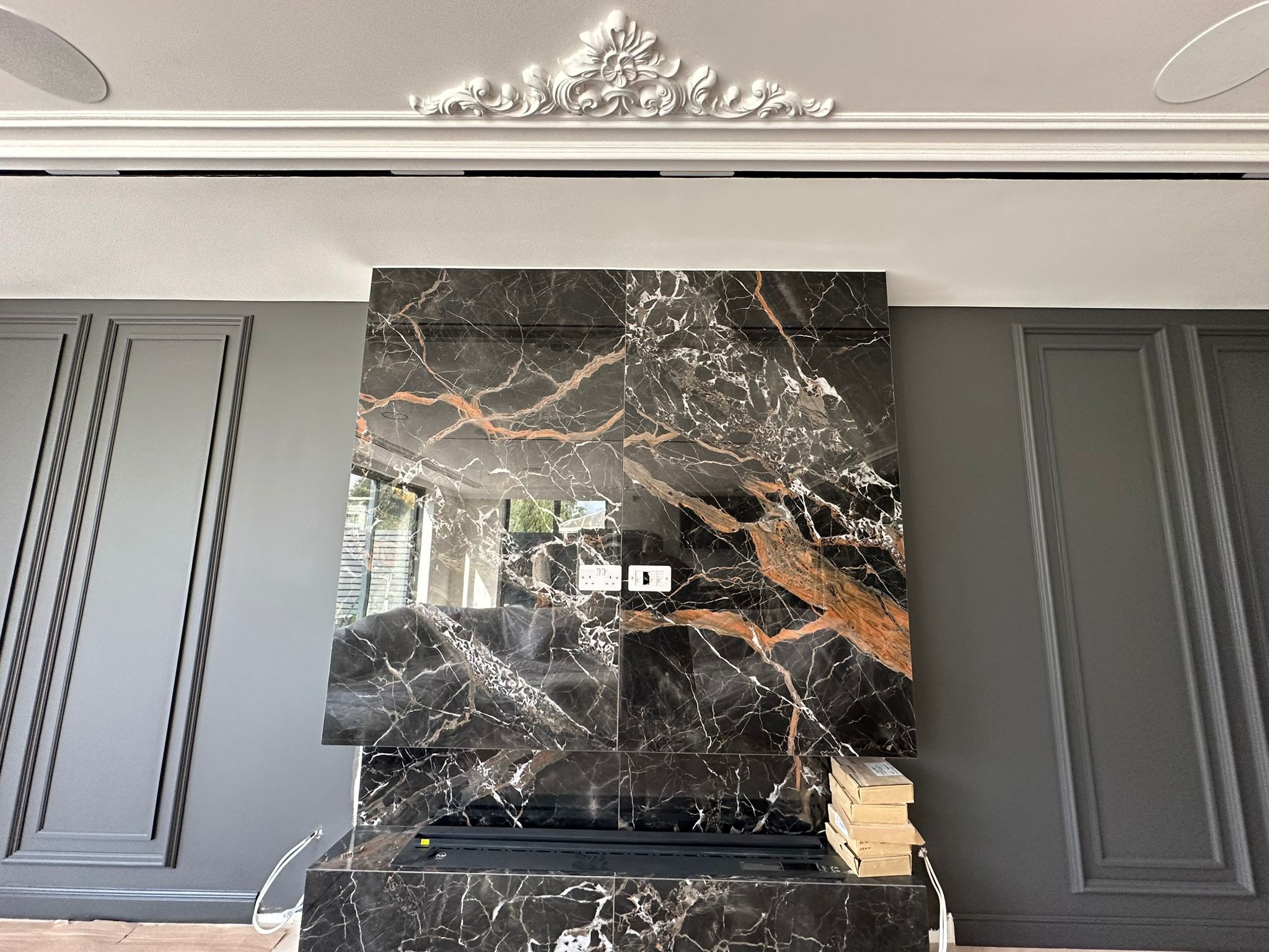 Elegancki fragment salonu z marmurową ścianą, dekoracyjnym gzymsem i szarą boazerią. Widoczne gniazdka elektryczne i elementy montażowe.