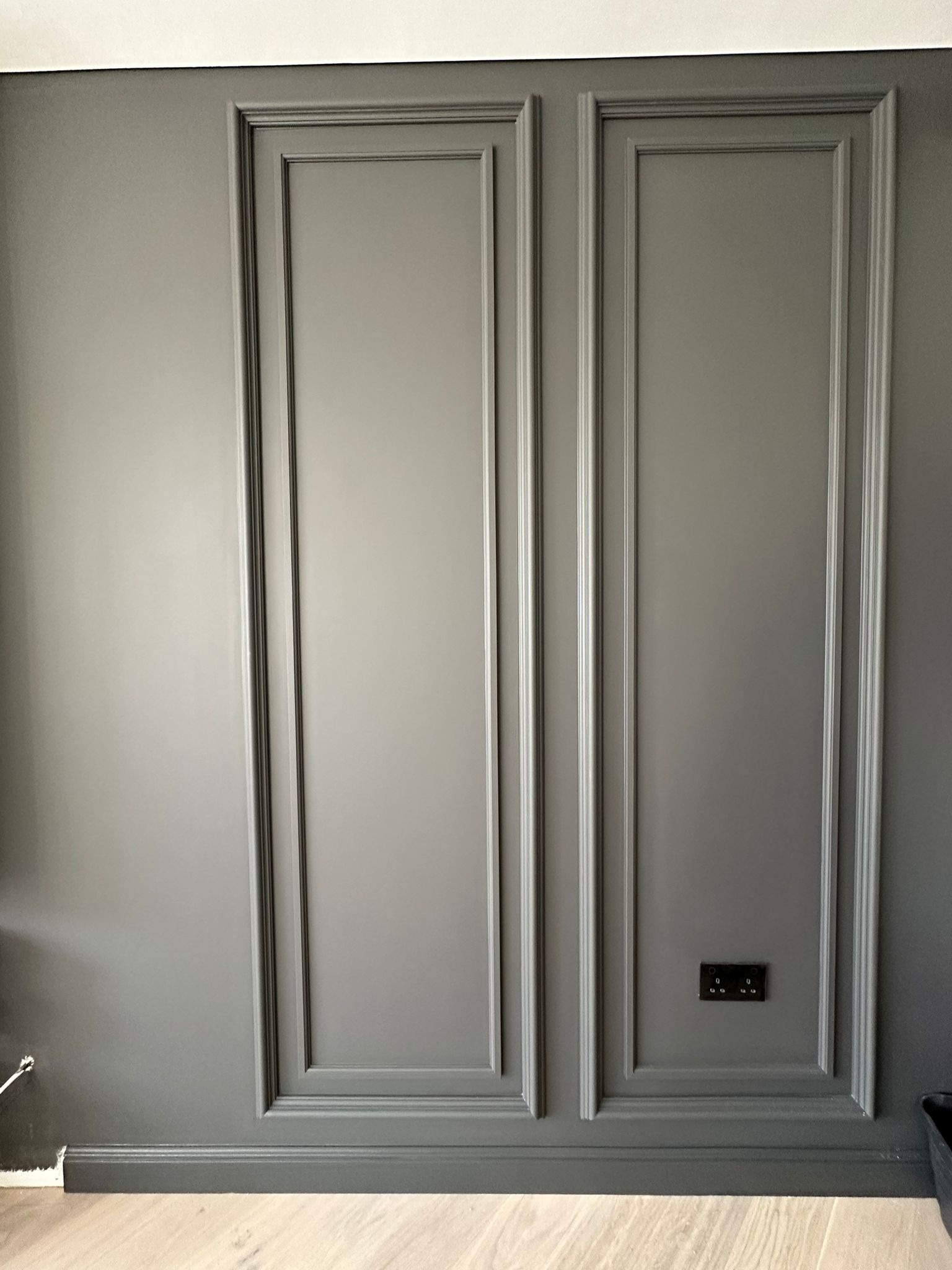 Szara ściana z dekoracyjnymi panelami w stylu klasycznym i gniazdkiem elektrycznym. Podłoga drewniana. Wykończenie wnętrza w stonowanej kolorystyce.