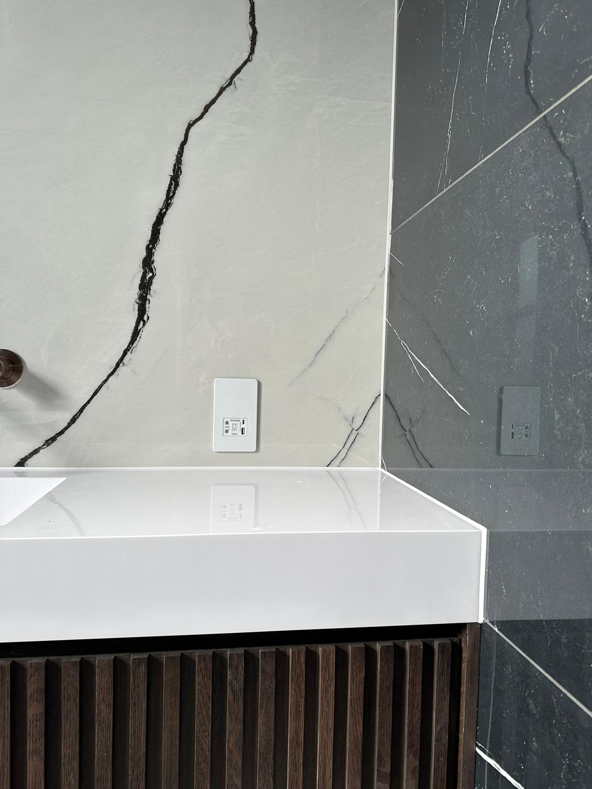 Nowoczesna umywalka z szafką z ciemnego drewna na tle płytek imitujących marmur w odcieniach szarości i bieli, widoczne detale wykończenia.