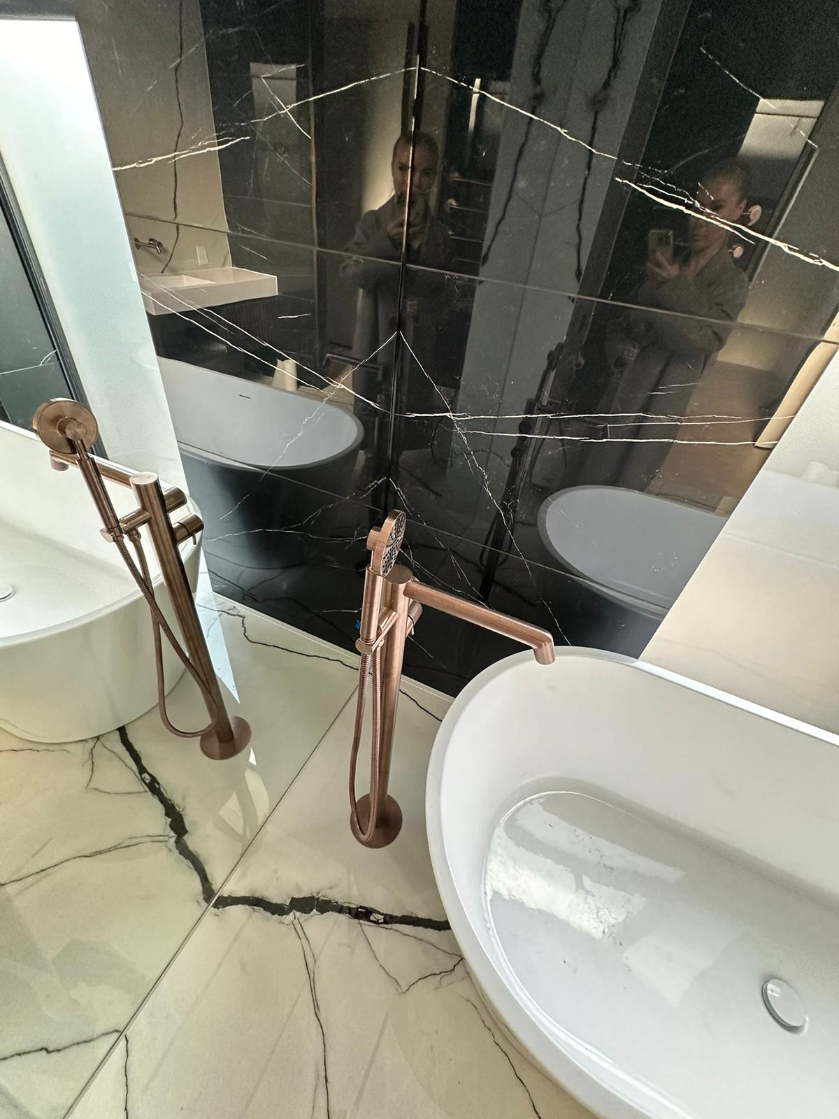 Ekskluzywna łazienka z czarnym marmurem, owalną wanną i miedzianą armaturą. Widoczne odbicie osoby robiącej zdjęcie w lustrzanej ścianie.