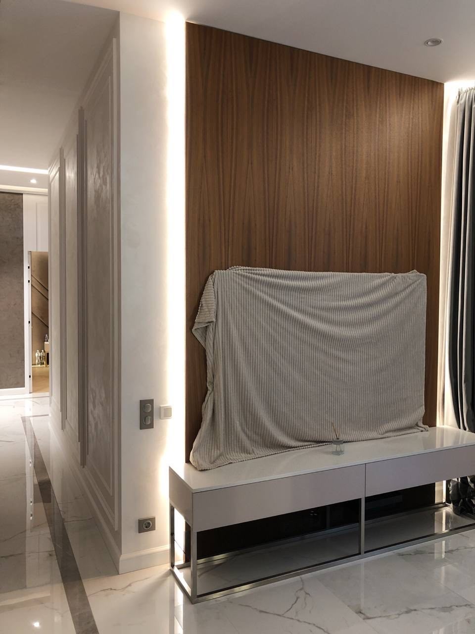 Nowoczesny salon z drewnianą ścianą akcentującą, oświetleniem LED i marmurową podłogą. Telewizor przykryty pledem, minimalistyczna szafka RTV.