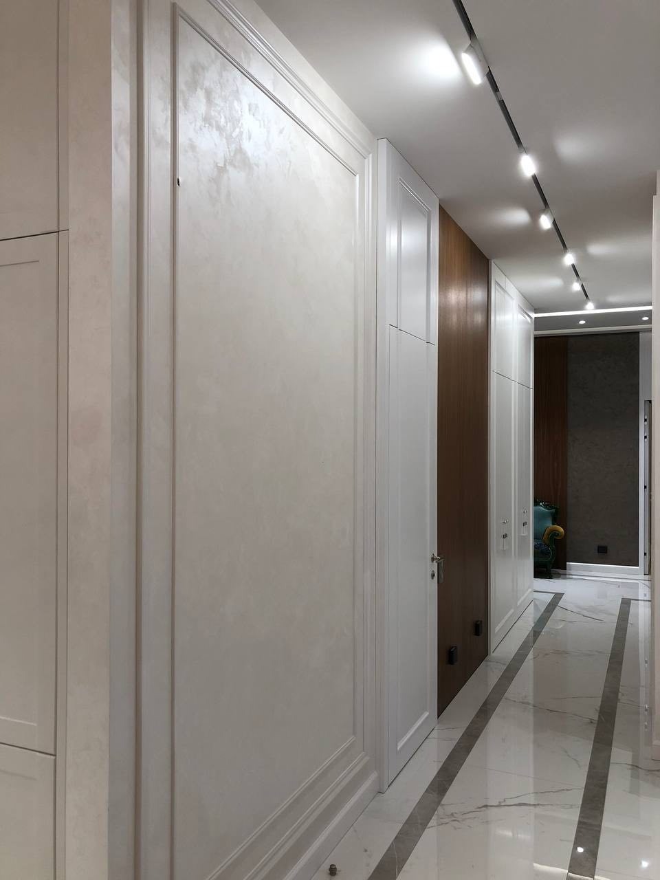 Korytarz w neutralnej tonacji z marmurową podłogą i oświetleniem szynowym. Ściany wykończone w odcieniach bieli z dekoracyjnymi panelami. Widoczne drzwi w zabudowie.