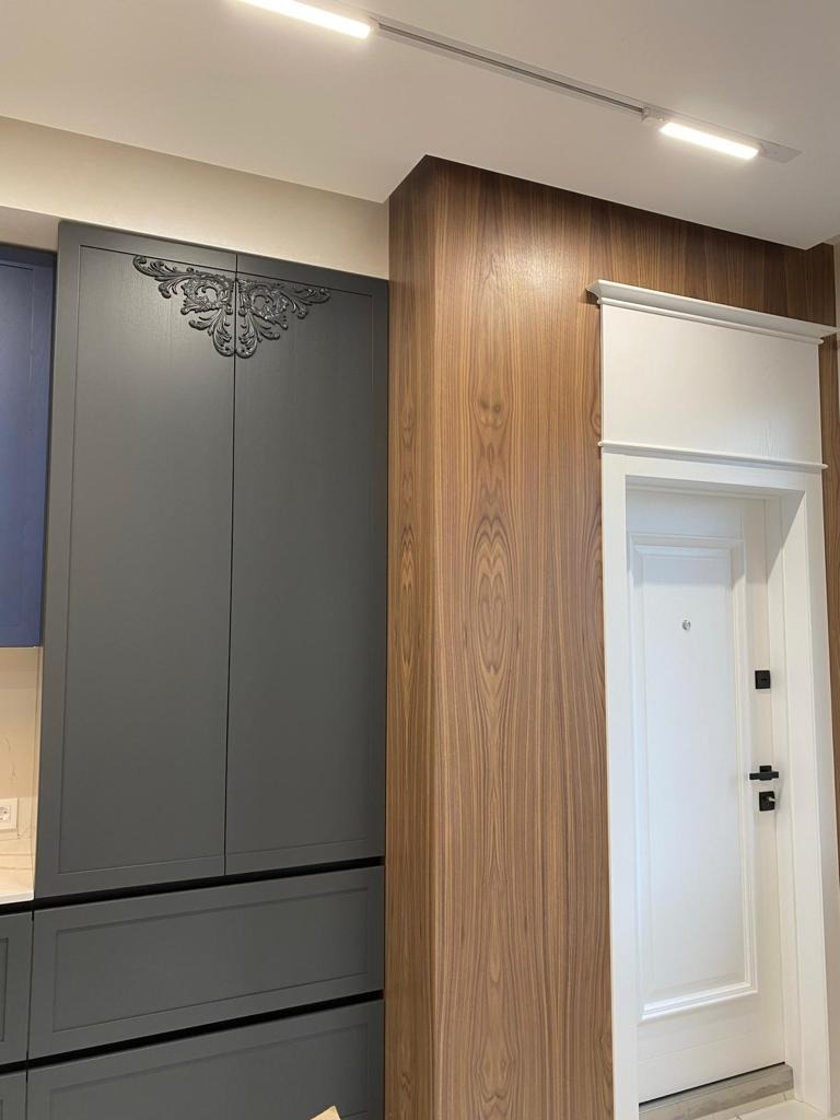 Szara zabudowa kuchenna z dekoracyjnym ornamentem sąsiaduje z drewnianą okładziną ściany i białymi drzwiami wejściowymi. Oświetlenie punktowe sufitowe.