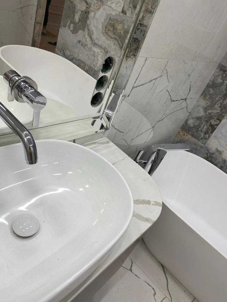 Nowoczesna łazienka: biała umywalka nablatowa, bateria chromowana, lustro z oświetleniem punktowym i marmurowe płytki ścienne tworzą elegancki wystrój.