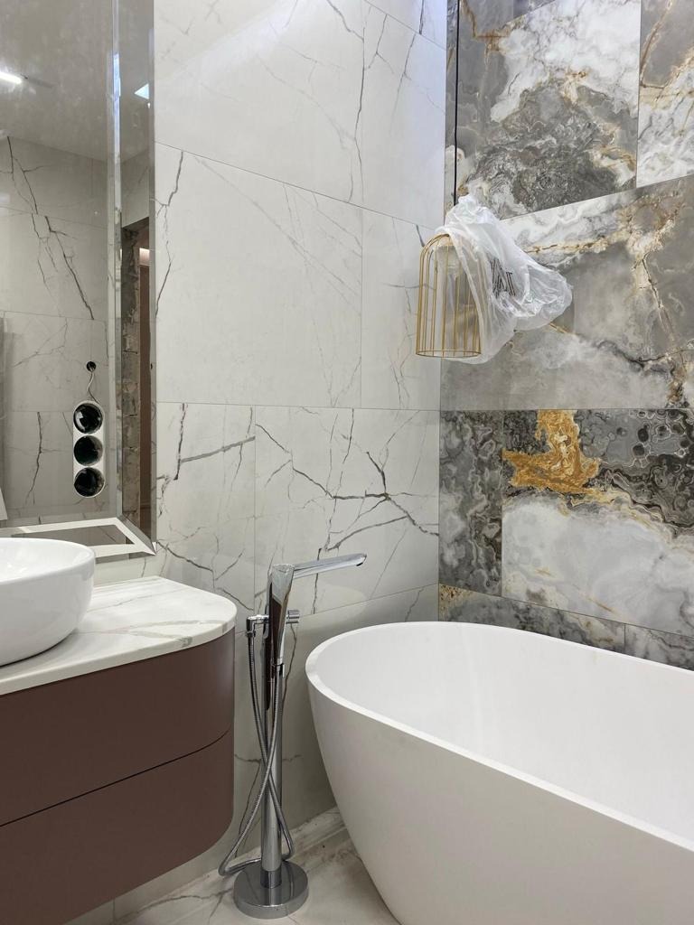 Nowoczesna łazienka z wolnostojącą wanną, marmurowymi płytkami na ścianach i wiszącą lampą w złotej klatce, jeszcze w folii ochronnej, obok umywalka na brązowej szafce.