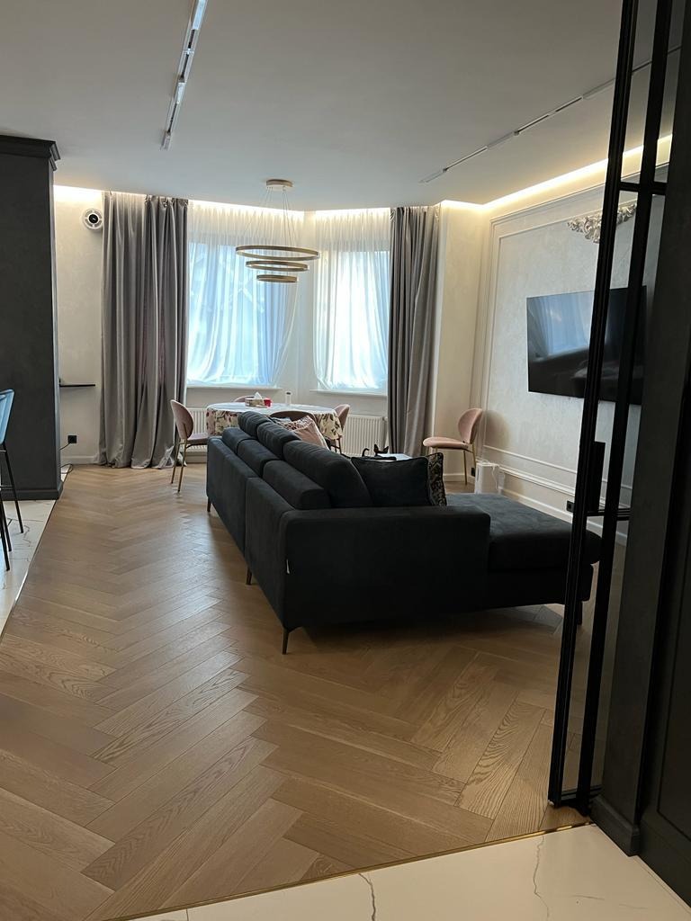 Elegancki salon z parkietem w jodełkę, ciemną sofą i dekoracyjnymi zasłonami. Nowoczesne oświetlenie i telewizor na ścianie. Stonowana kolorystyka, jasne ściany.