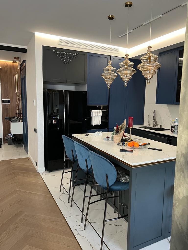 Elegancka kuchnia z wyspą, marmurowym blatem i niebieskimi szafkami. Nad wyspą wiszą trzy ozdobne, szklane lampy. Podłoga wyłożona parkietem w jodełkę.