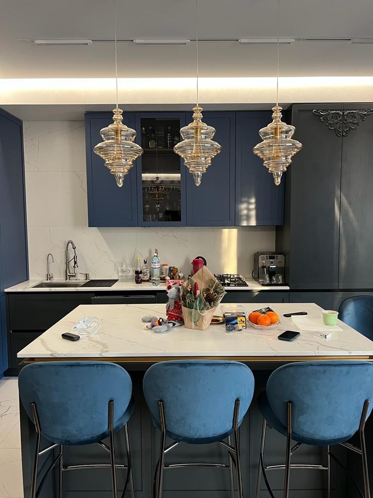 Nowoczesna kuchnia w odcieniach granatu i bieli z marmurowym blatem wyspowym, oświetlona trzema designerskimi lampami wiszącymi. Widoczne detale i dodatki.