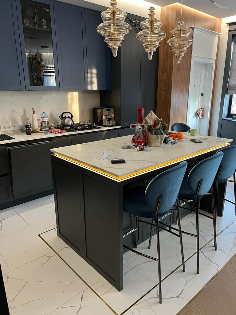 Niebieska kuchnia z wyspą oświetloną listwą LED i marmurowym blatem. Nad wyspą wiszą trzy szklane lampy. Marmurowe płytki na podłodze z akcentami w kolorze złotym.