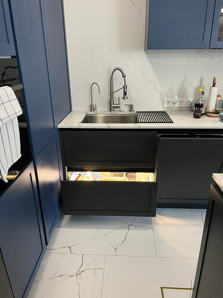 Nowoczesna kuchnia z granatowymi szafkami i marmurowym blatem. Otwarta szuflada pod zlewem z oświetleniem LED. Bateria kuchenna ze stali nierdzewnej.