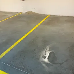 Szary beton z żółtymi liniami w hali. Widoczny fragment instalacji przy ścianie i ślady po niedawnym czyszczeniu podłoża, które odbija światło.