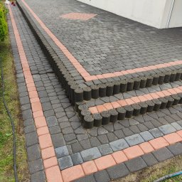 CLEAN HOUSE Radosław Myśliwiec - Ułożona kostka brukowa w odcieniach szarości i czerwieni, tworząca schody i podjazd. Widoczne cylindryczne obrzeża i fragment budynku w tle.