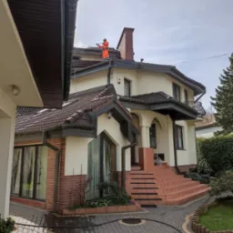 Pracownik w pomarańczowym kombinezonie myje dach domu z ceglanym kominem za pomocą myjki ciśnieniowej. Na kostce brukowej widać ślady wody.
