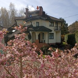 CLEAN HOUSE Radosław Myśliwiec - Pracownik w pomarańczowym kombinezonie czyści dach domu myjką ciśnieniową, na pierwszym planie kwitnąca magnolia.
