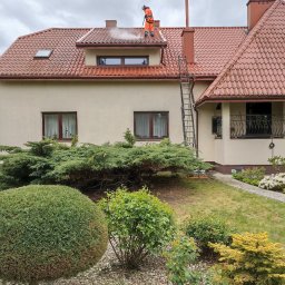 CLEAN HOUSE Radosław Myśliwiec - Pracownik w kombinezonie myje dach z czerwonej dachówki domu jednorodzinnego za pomocą myjki ciśnieniowej. Widoczna drabina oparta o budynek i zadbany ogród.