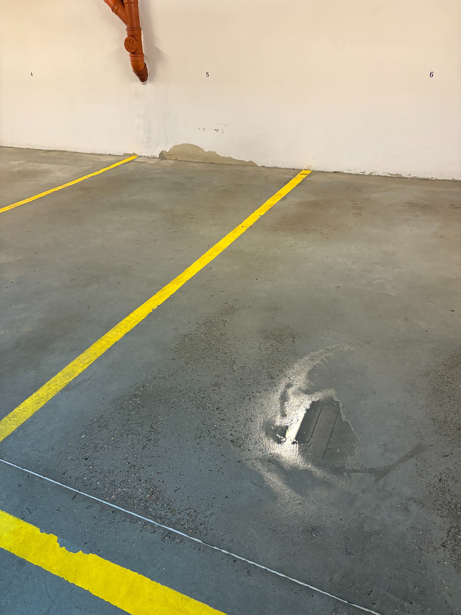 Szary beton z żółtymi liniami w hali. Widoczny fragment instalacji przy ścianie i ślady po niedawnym czyszczeniu podłoża, które odbija światło.