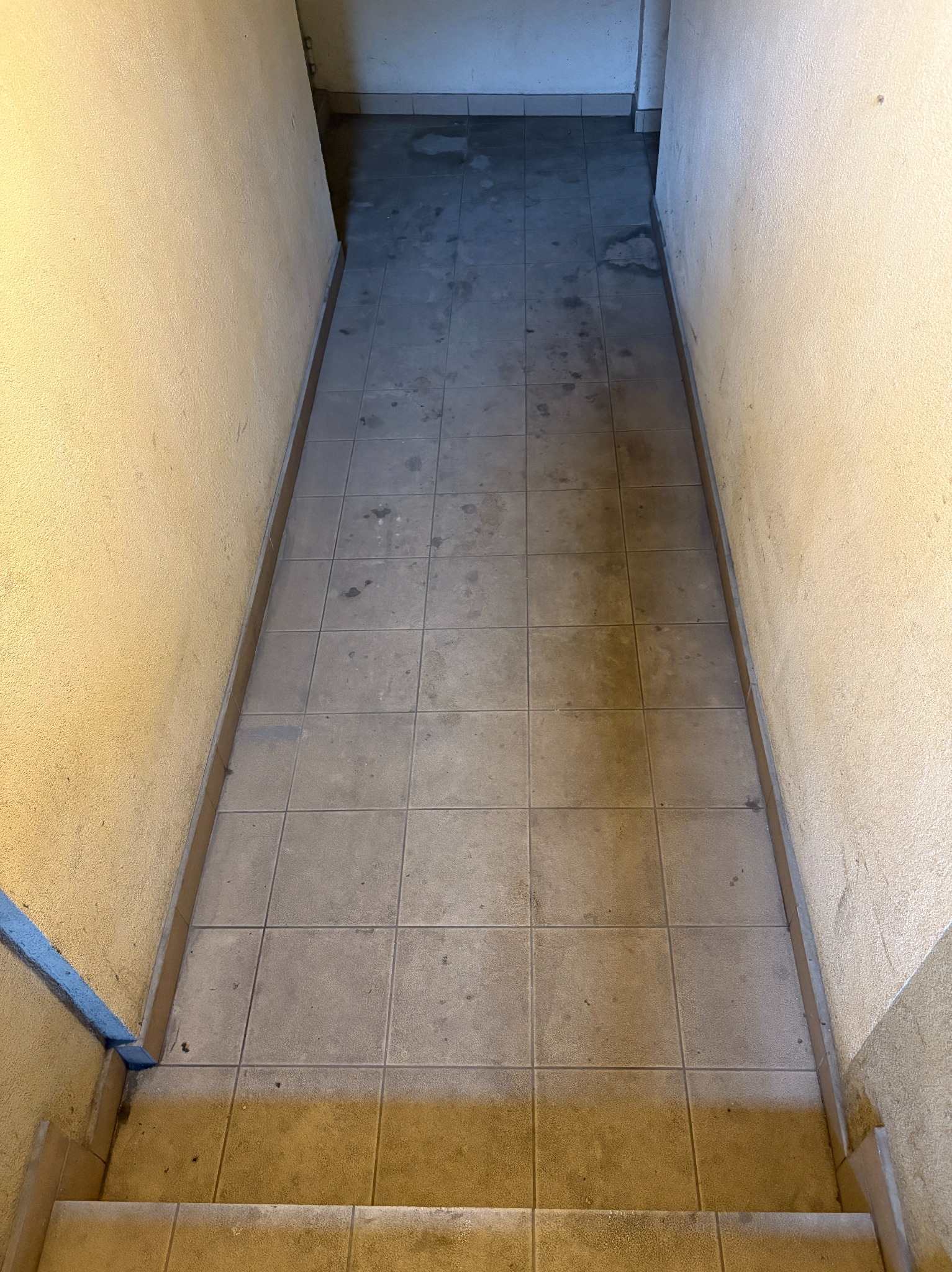 Brudna podłoga z płytek w wąskim korytarzu z żółtymi ścianami. Widoczne plamy i zabrudzenia na podłodze. Perspektywa z góry, schody na pierwszym planie.