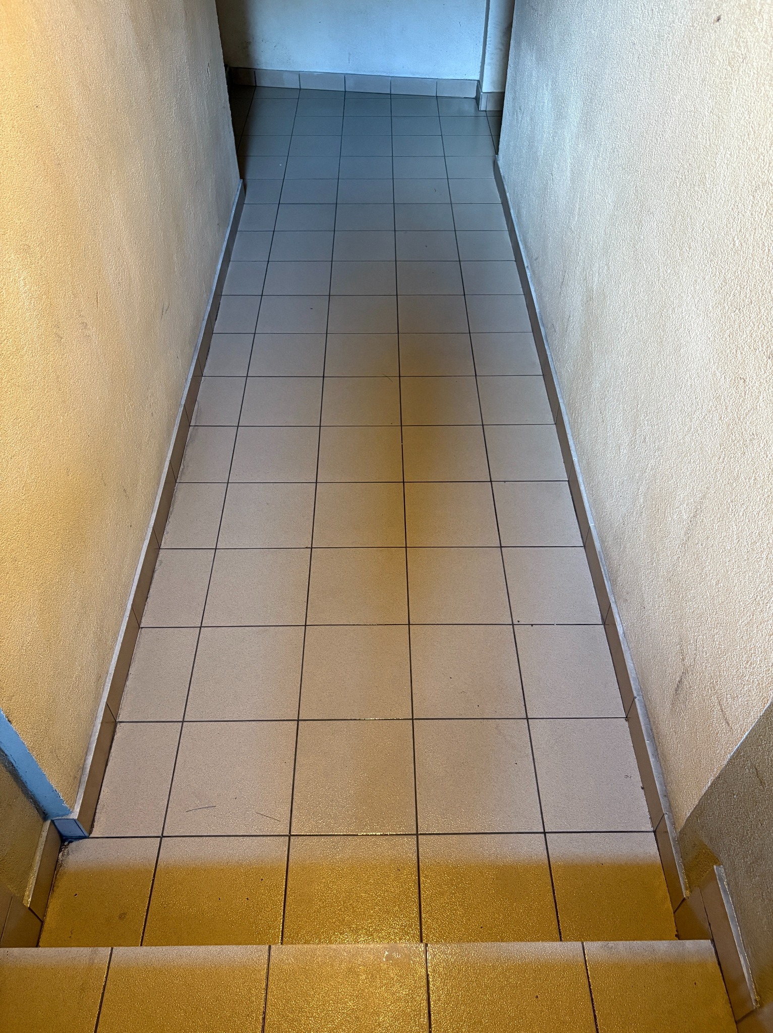 Kafelkowane schody w dół, kremowe ściany z wyraźną fakturą. Widoczne zabrudzenia na fugach i płytkach. Oświetlenie od góry podkreśla geometryczny układ.