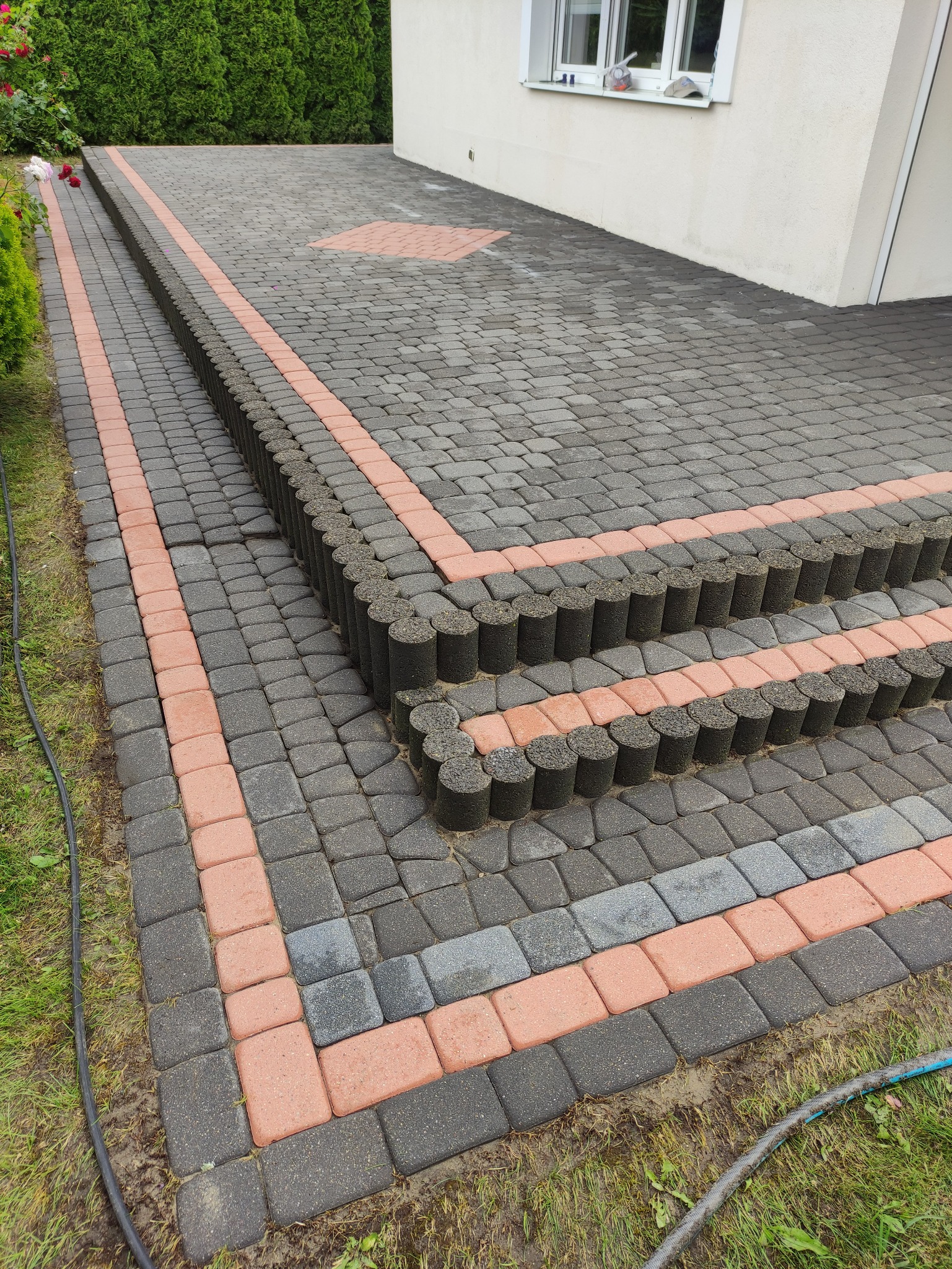 Ułożona kostka brukowa w odcieniach szarości i czerwieni, tworząca schody i podjazd. Widoczne cylindryczne obrzeża i fragment budynku w tle.