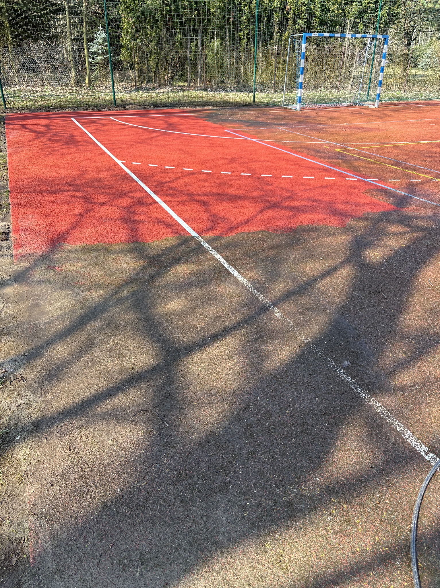 Czerwone boisko sportowe z bramką w tle, widoczne cienie drzew. Część boiska jest wyraźnie czystsza od reszty, co sugeruje niedawne mycie.