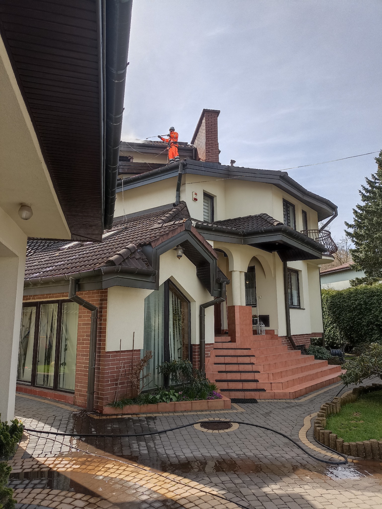 Pracownik w pomarańczowym kombinezonie myje dach domu z ceglanym kominem za pomocą myjki ciśnieniowej. Na kostce brukowej widać ślady wody.